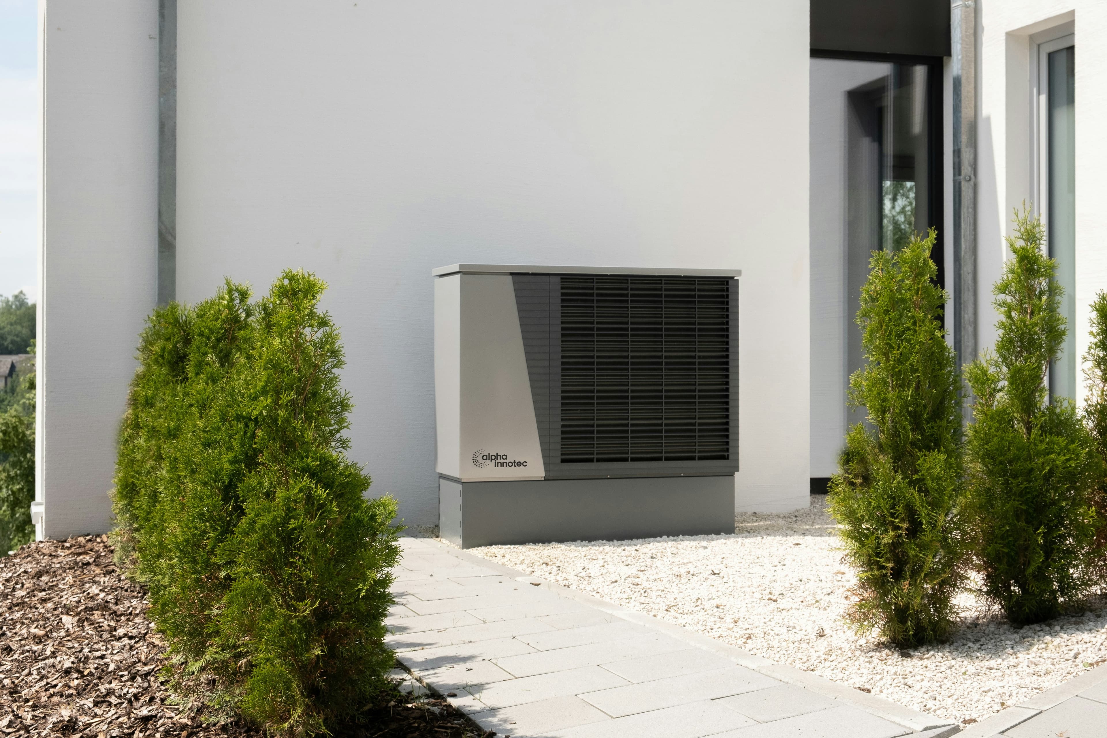 Grey alpha innotec heat pump unit installed outside a white house next to green bushes.