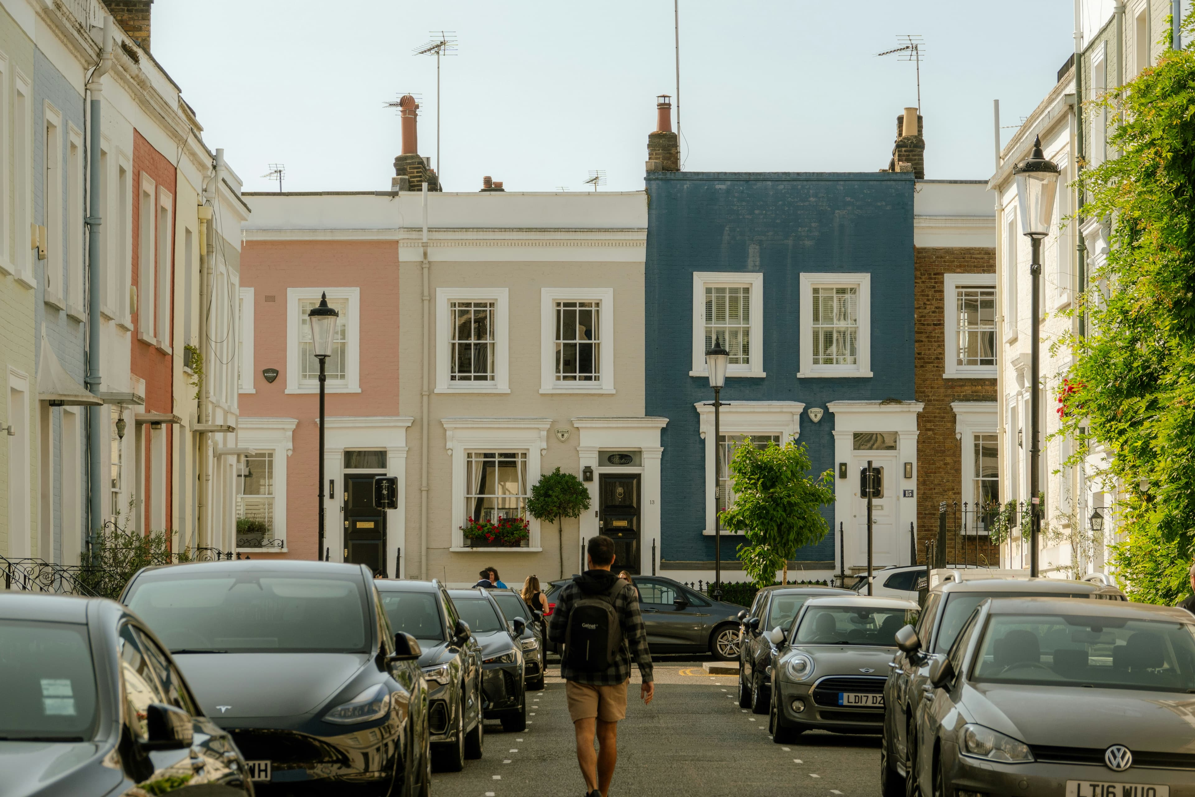 A man walks down a narrow street lined with colorful terraced houses and parked cars.