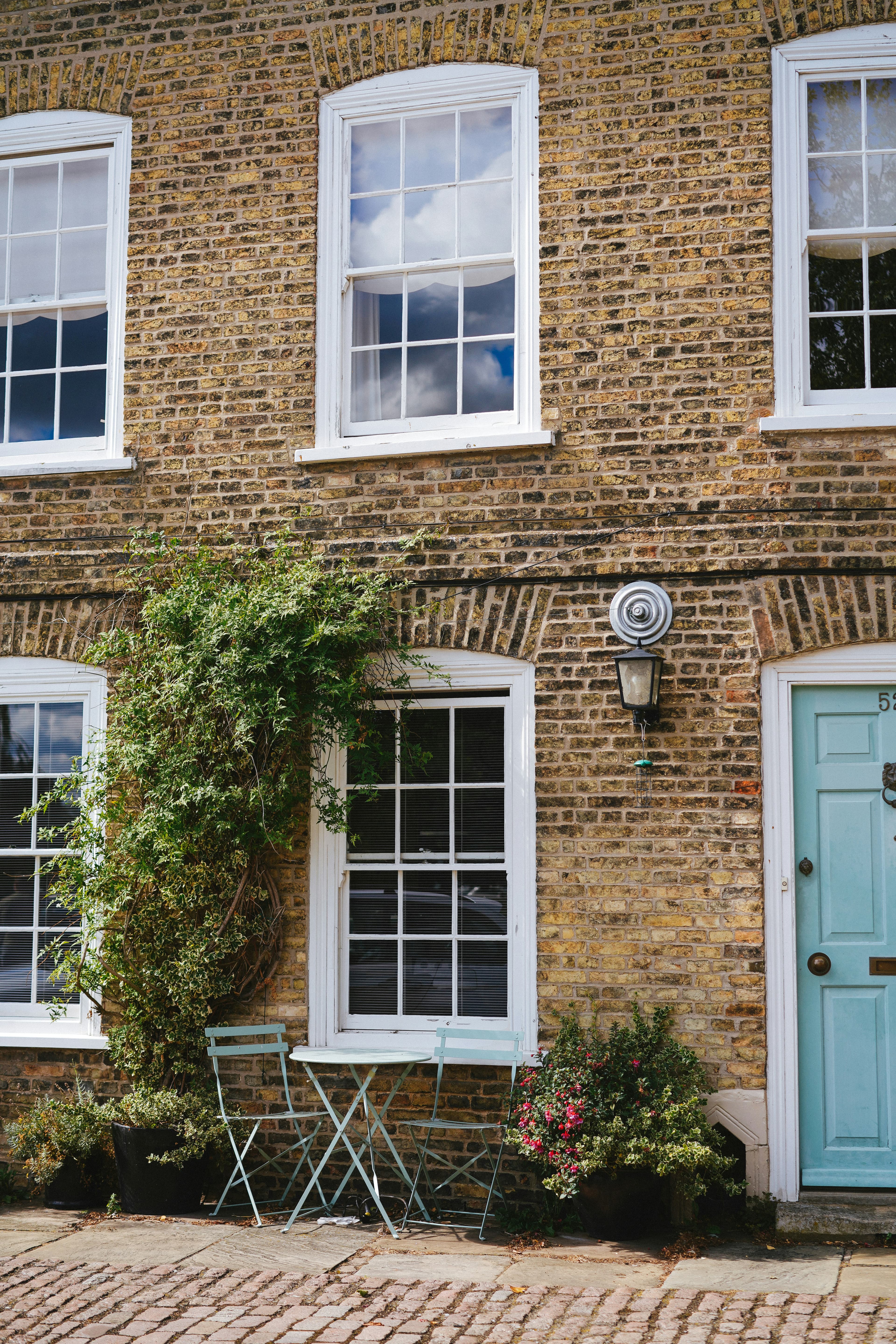 Tan brick building facade with white windows, a light blue door, and bistro seating.
