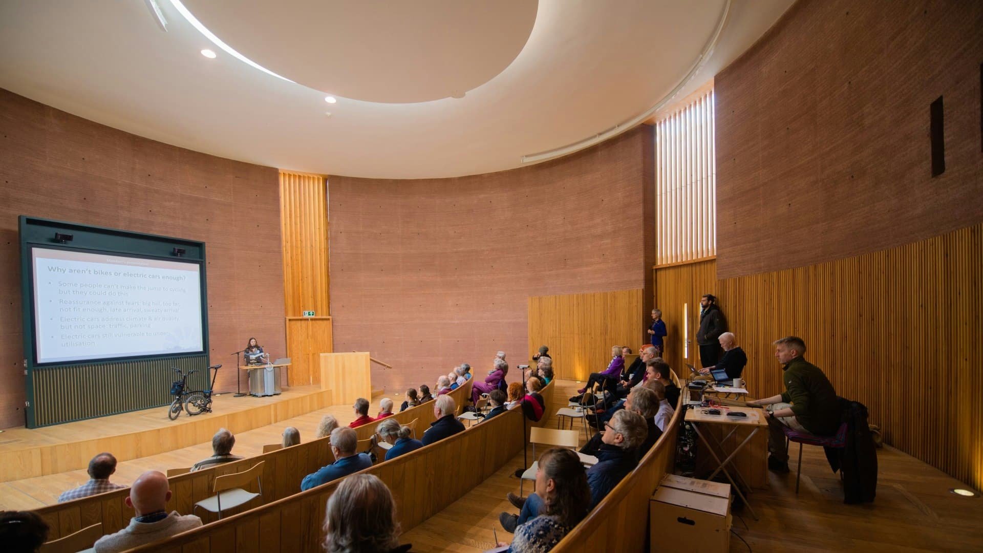 Audience in a circular wooden auditorium watching a presentation about electric vehicles and bicycles.